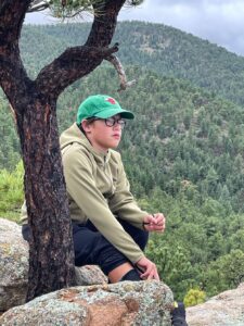 a young student wearing a green hat kneeling by a tree trunk with mountains in the background